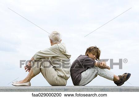 Stock Image - Grandmother and grandson sitting back to back, woman looking away, boy looking at camera. Fotosearch