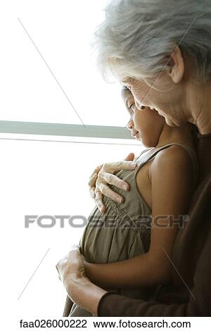 Stock Image - Grandmother hugging granddaughter, cheek to cheek, both looking out window. Fotosearch