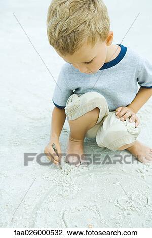 Stock Image - Little boy crouching at the beach, drawing in sand with stick. Fotosearch