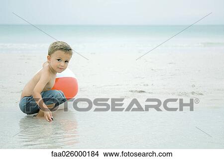 Little boy crouching in water at the beach, playing with beach ball, looking at camera View Large Photo Image Picture - Little boy crouching in water at the beach, playing with beach ball, looking at camera. Fotosearch