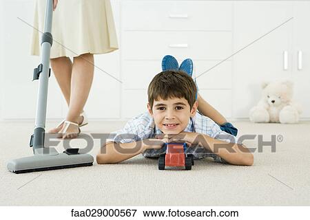 Stock Photo - Little boy lying on the ground with toys, smiling at camera, mother vacuuming around him. Fotosearch