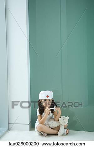 Stock Image - Little girl wearing nurse's hat, sitting on the ground with teddy bear on lap. Fotosearch