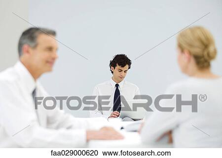 Stock Image - Male professional using laptop computer, colleagues talking in foreground. Fotosearch