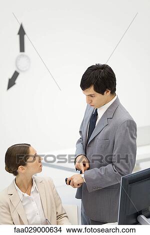 Male supervisor standing beside female employee, pointing at wristwatch, clock hands on wall in background View Large Photo Image Picture - Male supervisor standing beside female employee, pointing at wristwatch, clock hands on wall in background. Fotosearch