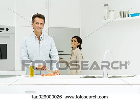 Man and woman in kitchen, man chopping tomatoes while woman goes toward refrigerator, both smiling at camera View Large Photo Image Stock Photograph - Man and woman in kitchen, man chopping tomatoes while woman goes toward refrigerator, both smiling at camera. Fotosearch