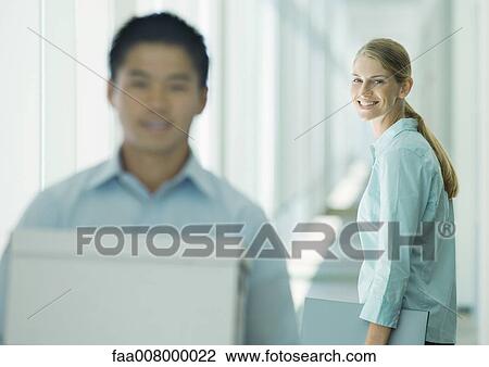 Stock Image - Man carrying boxes in hallway, female colleague watching. Fotosearch