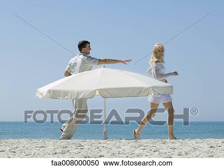 Stock Image - Man chasing woman around parasol on beach. Fotosearch