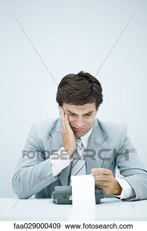 Stock Photo - Man in suit looking at printout on calculator, holding head. Fotosearch
