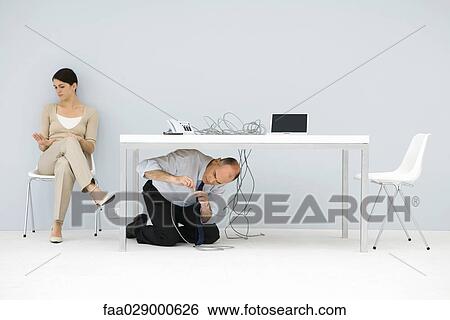 Man kneeling under desk, connecting tangled wires, woman sitting in chair, looking at hand View Large Photo Image Stock Photograph - Man kneeling under desk, connecting tangled wires, woman sitting in chair, looking at hand. Fotosearch