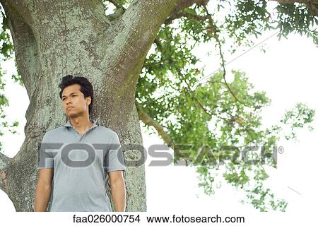 Picture - Man leaning against tree, looking away, waist up. Fotosearch