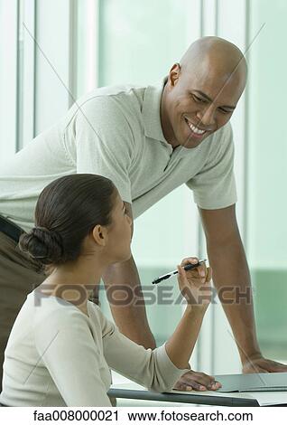 Stock Image - Man leaning on desk next to female coworker, smiling. Fotosearch