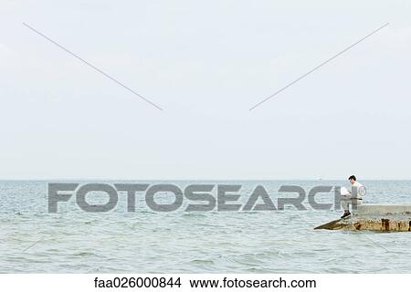 Picture - Man sitting at the end of pier, reading book, in the distance. Fotosearch