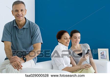 Stock Photo - Man sitting on sofa, smiling at camera, his wife and daughter looking at cell phone behind him. Fotosearch