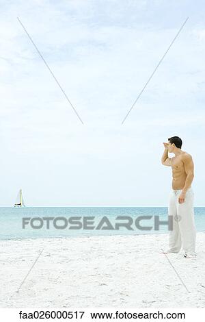 Man standing at the beach, looking at sailboat in the distance, side view View Large Photo Image Stock Photo - Man standing at the beach, looking at sailboat in the distance, side view. Fotosearch