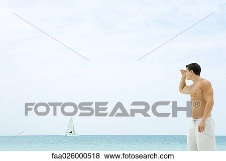 Man standing at the beach, looking at view, sailboat in the distance View Large Photo Image Stock Photo - Man standing at the beach, looking at view, sailboat in the distance. Fotosearch