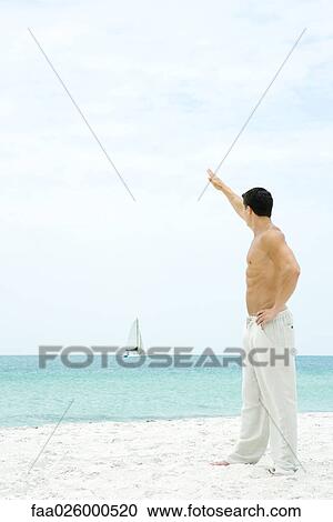 Man standing at the beach with arm raised, waving at sailboat in the distance, side view View Large Photo Image Stock Image - Man standing at the beach with arm raised, waving at sailboat in the distance, side view. Fotosearch