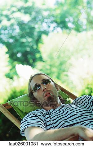 Stock Image - Man wearing sunglasses reclining in chair, smoking hand-rolled cigarette. Fotosearch