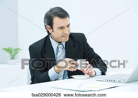 Stock Photograph - Mature businessman sitting at desk with cup of coffee, smiling. Fotosearch