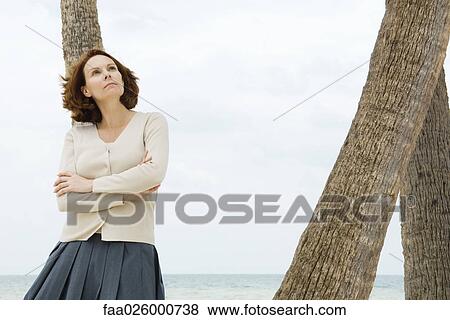 Mature woman leaning against palm tree with arms folded, looking up View Large Photo Image Stock Photo - Mature woman leaning against palm tree with arms folded, looking up. Fotosearch