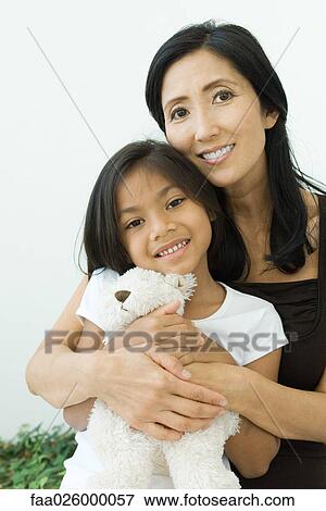 Stock Photo - Mother and daughter embracing teddy bear together, both smiling at camera, portrait. Fotosearch
