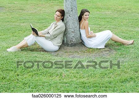 Mother and daughter leaning against tree trunk, woman reading book, teen girl smiling at camera View Large Photo Image Stock Photography - Mother and daughter leaning against tree trunk, woman reading book, teen girl smiling at camera. Fotosearch