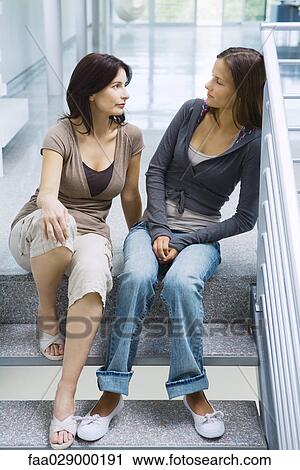 Stock Image - Mother and daughter sitting together on stairs, looking at each other, full length. Fotosearch