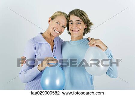 Stock Image - Mother and teen son with arms around each other's shoulders, holding balloons, smiling at camera. Fotosearch