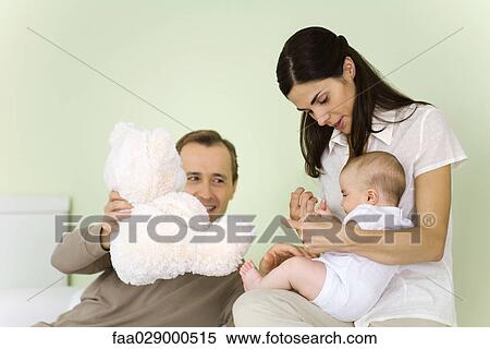 Stock Photography - Mother holding infant on lap, father holding teddy bear, smiling. Fotosearch