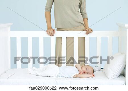 Stock Image - Mother standing behind crib as infant sleeps, cropped view. Fotosearch