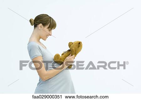 Stock Image - Pregnant woman holding teddy bear, smiling, side view. Fotosearch