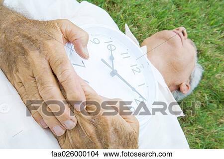 Senior man lying on grass, holding clock, cropped view View Large Photo Image Picture - Senior man lying on grass, holding clock, cropped view. Fotosearch