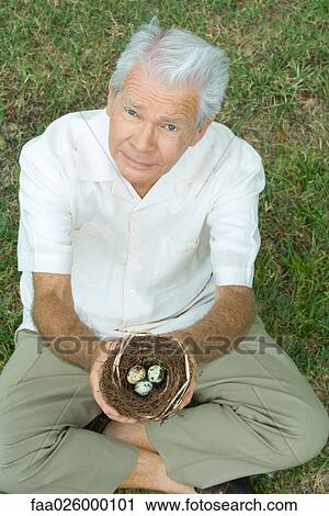 Stock Image - Senior man sitting on the ground, holding bird's nest with eggs, looking up at camera. Fotosearch