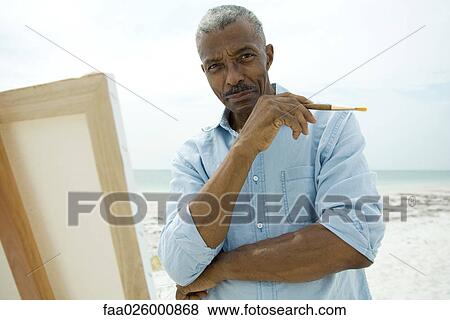 Stock Photo - Senior man standing in front of canvas at the beach, holding paint brush, smiling at camera. Fotosearch