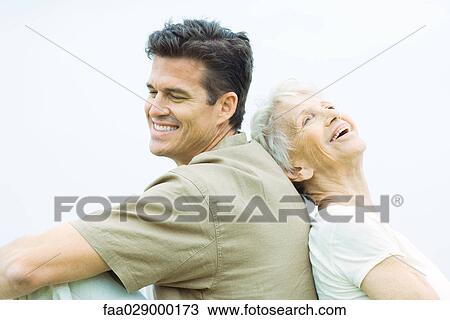 Stock Image - Senior woman and adult son sitting back to back, smiling, side view. Fotosearch