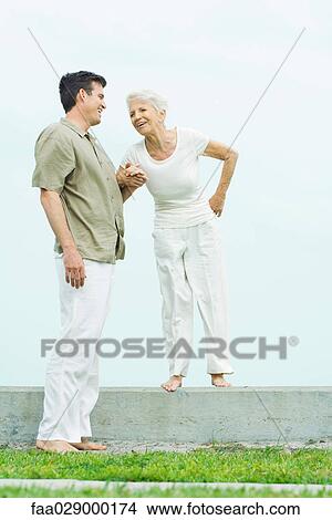 Picture - Senior woman standing on low wall next to adult son, holding hands, smiling. Fotosearch