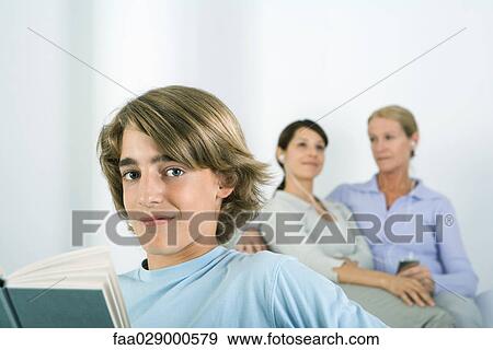 Stock Photo - Teenage boy holding book, smiling at camera, women listening to MP3 player in background. Fotosearch