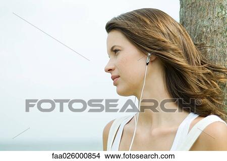 Picture - Teenage girl listening to earphones outdoors, hair tousled by breeze, looking away. Fotosearch