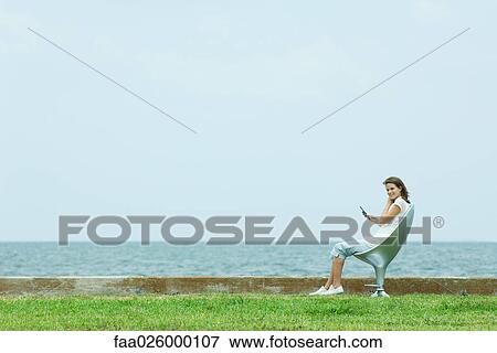 Stock Photo - Teenage girl sitting in chair by the sea, holding cell phone. Fotosearch
