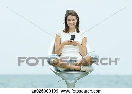 Teenage girl sitting in chair outdoors, holding cell phone, smiling at camera View Large Photo Image Stock Photo - Teenage girl sitting in chair outdoors, holding cell phone, smiling at camera. Fotosearch