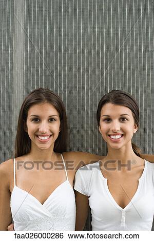 Stock Photograph - Teenage twin sisters with arms around one another, both smiling at camera, portrait. Fotosearch