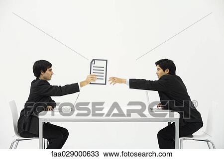 Stock Image - Two business associates sitting face to face at desk, one hand document to the other. Fotosearch