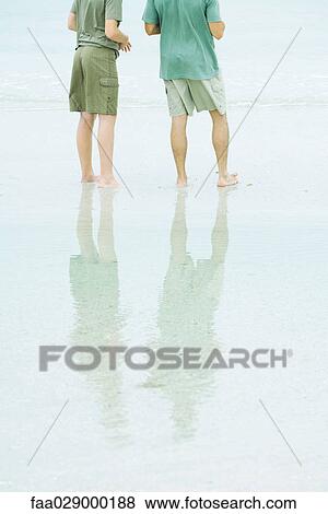 Stock Photo - Two males standing at the beach, rear view, cropped. Fotosearch