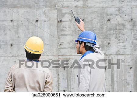 Two men in hard hats looking at cement wall, one holding up walkie-talkie View Large Photo Image Stock Image - Two men in hard hats looking at cement wall, one holding up walkie-talkie. Fotosearch