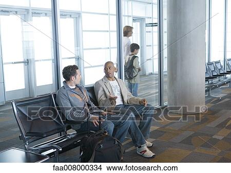 Two men sitting in airport lounge, travelers walking by in background View Large Photo Image Picture - Two men sitting in airport lounge, travelers walking by in background. Fotosearch