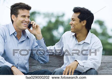Two men sitting on bench, smiling, one using cell phone View Large Photo Image Stock Image - Two men sitting on bench, smiling, one using cell phone. Fotosearch