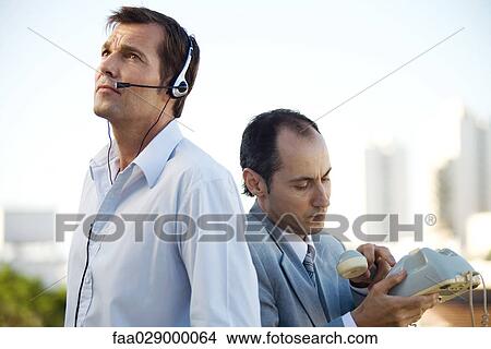 Two men standing back to back, one using headset, the other holding landline phone View Large Photo Image Picture - Two men standing back to back, one using headset, the other holding landline phone. Fotosearch