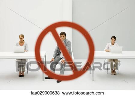Stock Photo - Two women seated in front of laptop computers, warning sign over man between them, man looking at camera. Fotosearch