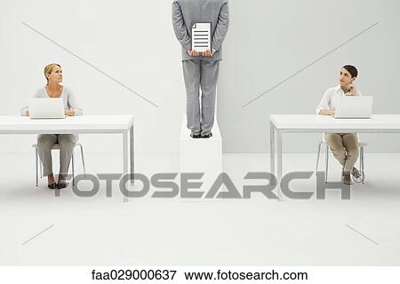 Two women sitting at desks with laptop computers, both looking at man holding document behind back View Large Photo Image Stock Photo - Two women sitting at desks with laptop computers, both looking at man holding document behind back. Fotosearch