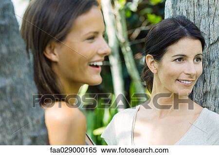 Stock Photography - Woman and teen girl leaning against tree trunks, looking away, close-up. Fotosearch