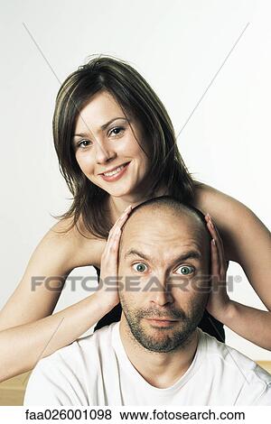 Stock Photo - Woman covering man's ears with her hands, smiling, both looking at camera. Fotosearch
